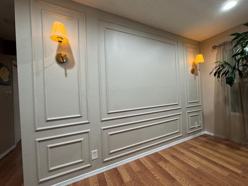 DIY wall molding kit installed in real home showing decorative wall panel result- Wall with decorative paneling, wall sconces, and a plant in a room.