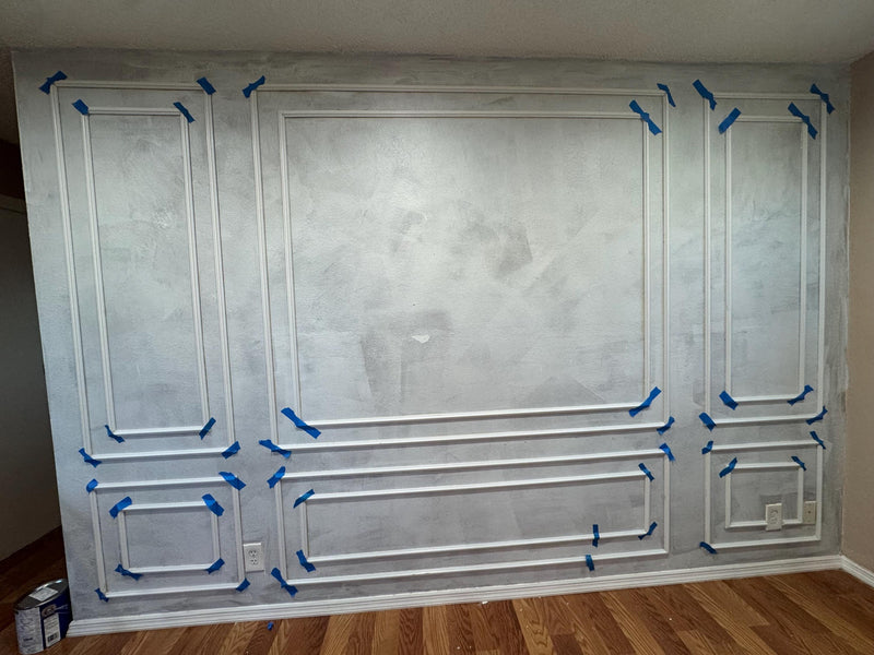 DIY wall molding kit installed in real home showing decorative wall panel result-Wall with decorative molding and blue tape, likely during renovation.