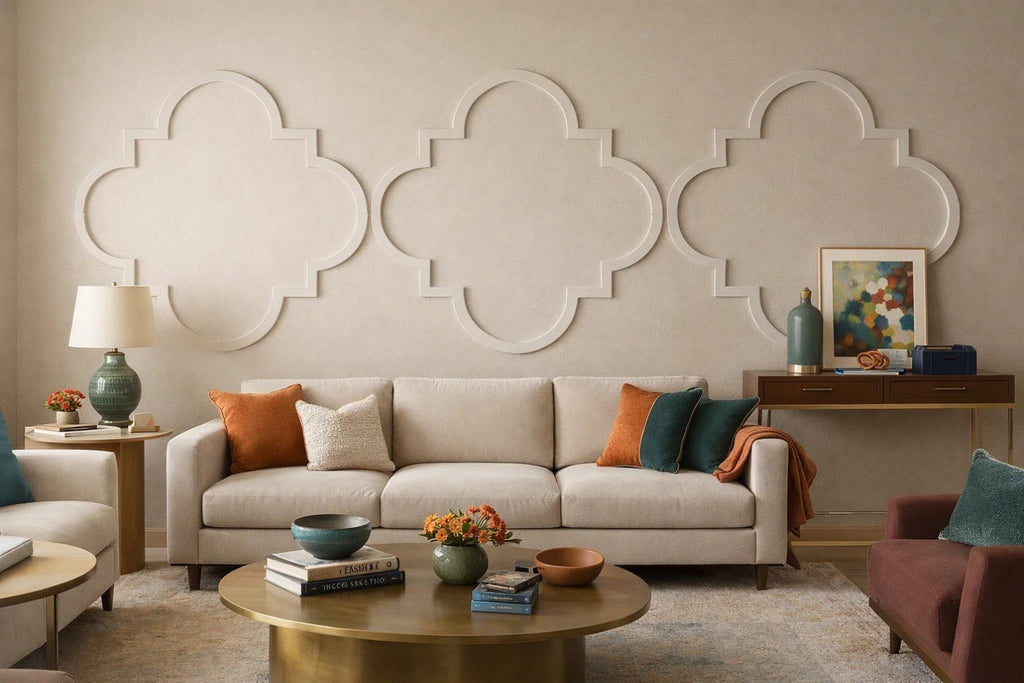 A styled living room featuring a beige fabric sofa with orange, cream, and teal accent pillows, set against a textured beige wall with three large white quatrefoil wall panels. A round gold coffee table, a green ceramic table lamp, and a dark wood console table with decorative items are arranged in front.